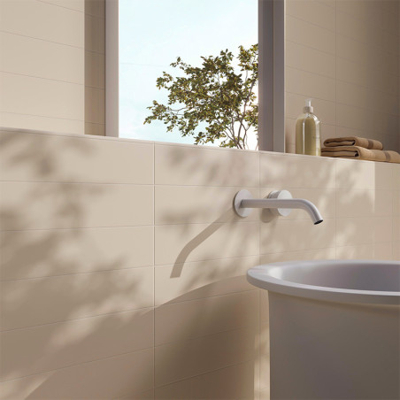 A bathtub faucet extends from a smooth, beige tiled wall. Sunlight streams through a window, casting tree shadows. A bottle and 