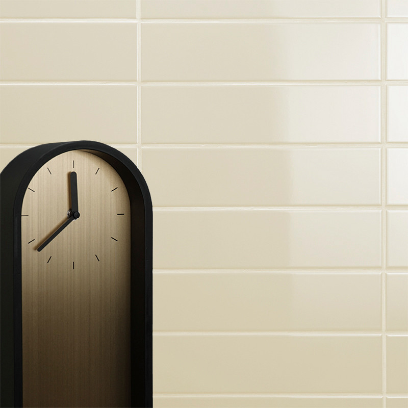 A minimalist black-framed clock displays no numbers, only black hour markings, against a beige tiled wall backdrop.
