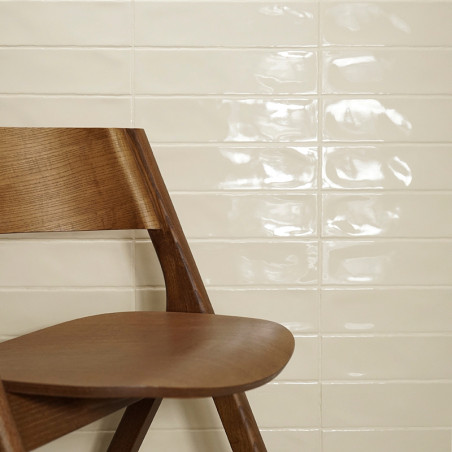 Ein Holzstuhl steht schräg vor einer glänzenden, cremefarbenen Fliesenwand, die das Licht reflektiert, in einem minimalistischen