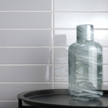 Une bouteille en verre tourbillonnante repose sur un plateau noir, adossé à un mur de carreaux de métro blanc brillant, reflétan