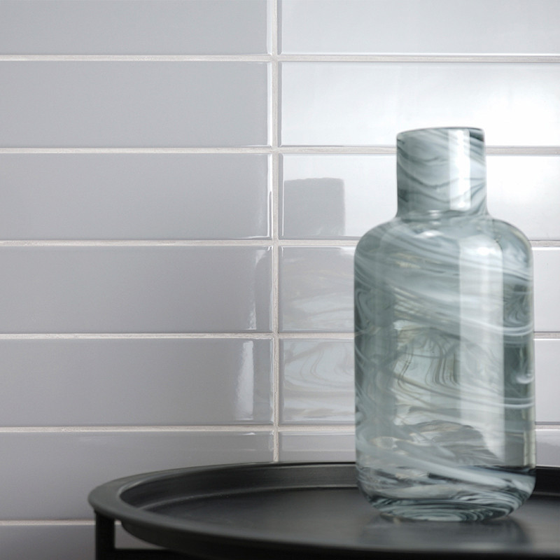 Een gewervelde glazen fles rust op een zwart dienblad, tegen een glanzende witte metrotegelmuur, en reflecteert het omgevingslic