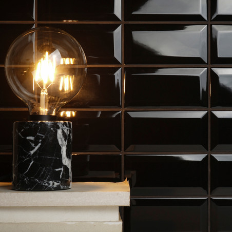 Eine leuchtende Glühbirne sitzt auf einem schwarzen Marmorsockel und beleuchtet einen glänzenden, schwarz gefliesten Wandhinterg