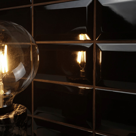 Eine leuchtende Glühbirne strahlt warmes Licht aus, das auf glänzend schwarzen, rechteckigen Fliesen reflektiert wird. Die Einri