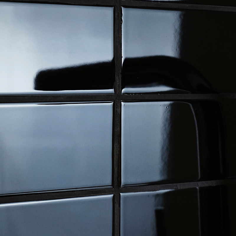 Ladrilhos cerâmicos pretos e brilhantes que refletem a luz formam um padrão de grade. Uma sombra se curva em um bloco, sugerindo