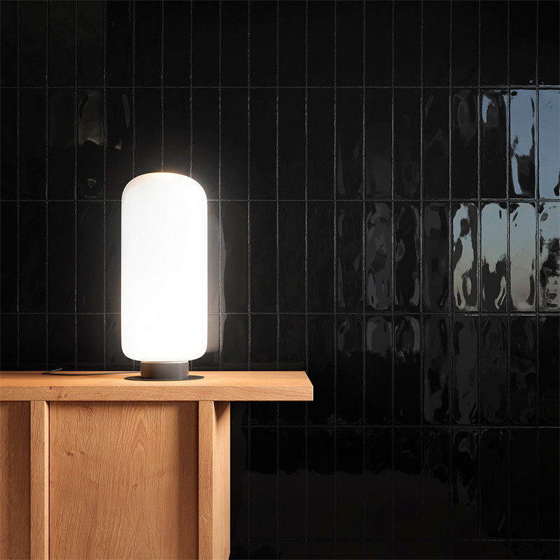 Uma lâmpada alta e cilíndrica emite luz suave sobre uma mesa de madeira, colocada contra uma parede de azulejos pretos brilhante