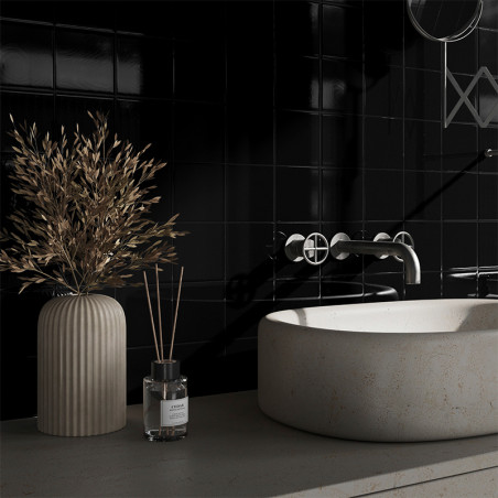 A round stone basin sits on a countertop beside a ribbed vase with dried branches. Black tiles cover the wall, and a sleek fauce