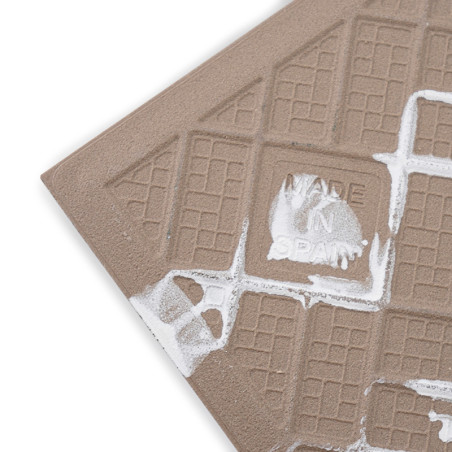 Een getextureerde bruine keramische tegel is geëtst met rasterpatronen en witte verfspatten, met de tekst `MADE IN SPAIN.`