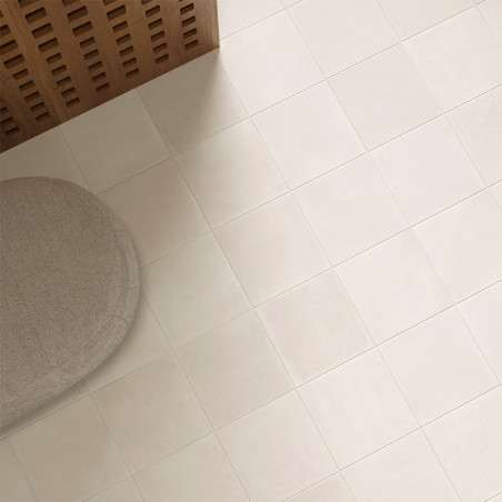 Ein hellbeiger Fliesenboden erstreckt sich über die gesamte Fläche. In einer Ecke neben einer Holzplatte steht ein runder Ottoma