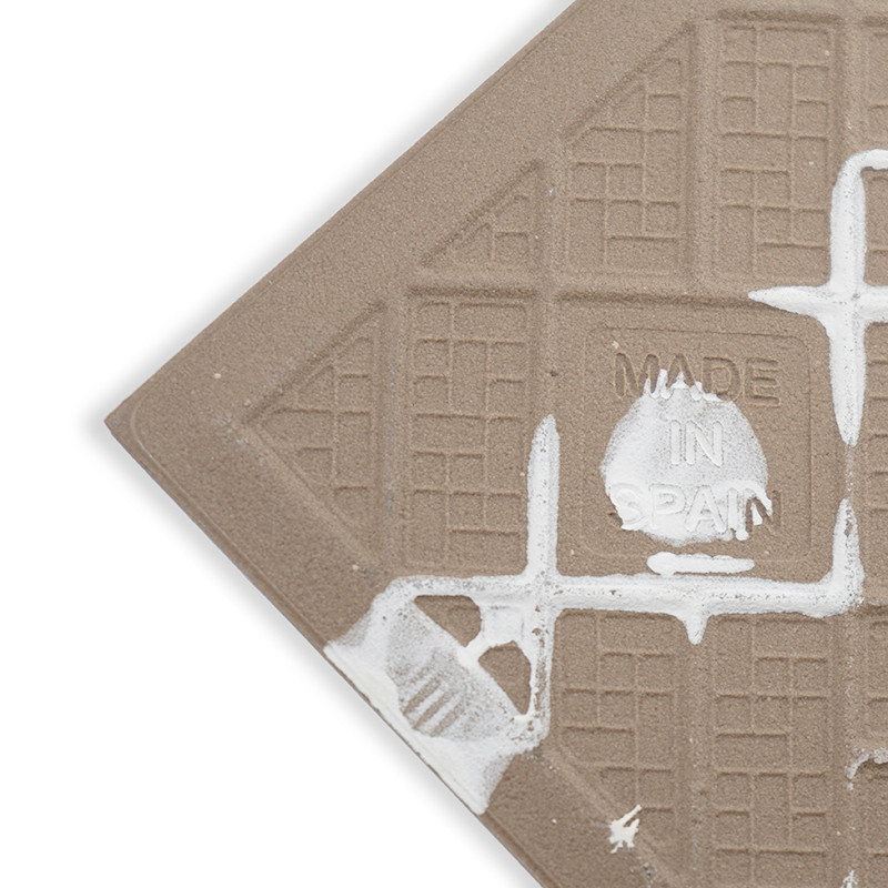 Un carreau de céramique texturé avec des motifs en grille présente des éclaboussures blanches. Le texte en relief indique « MADE