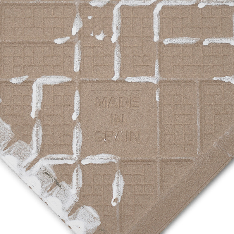 Un azulejo beige con la leyenda `MADE IN SPAIN` impreso y presenta un patrón de cuadrícula tipo gofre. Un residuo blanco texturi