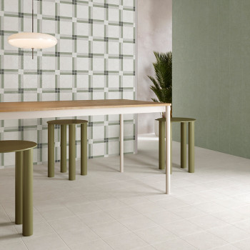 Een moderne eettafel staat op lichte vierkante tegels, omgeven door groene cilindervormige krukken. Een muur en plant met geruit