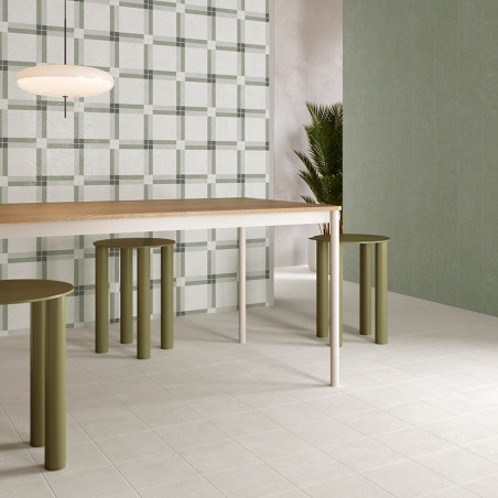 Een moderne eettafel staat op lichte vierkante tegels, omgeven door groene cilindervormige krukken. Een muur en plant met geruit