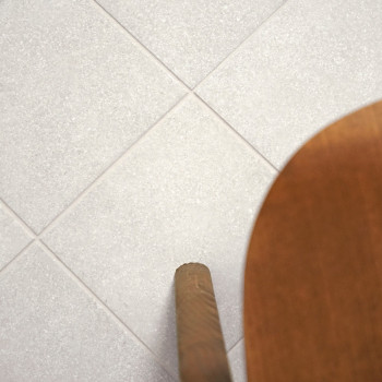 Een raster van lichtgrijze tegels ligt plat, met een houten stoelpoot die van rechts het frame binnenkomt, wat een binnenvloer s