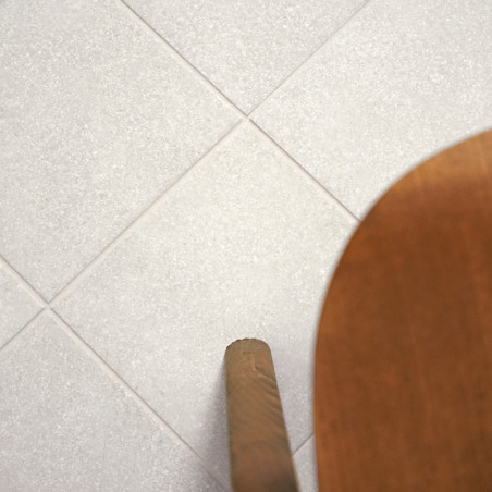 Een raster van lichtgrijze tegels ligt plat, met een houten stoelpoot die van rechts het frame binnenkomt, wat een binnenvloer s