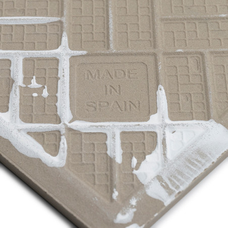 Um azulejo bege com padrões de grade em relevo apresenta o texto `MADE IN SPAIN`, parcialmente coberto por argamassa branca, con Um azulejo bege com padrões de grade em relevo apresenta o texto `MADE IN SPAIN`, parcialmente coberto por argamassa branca, con