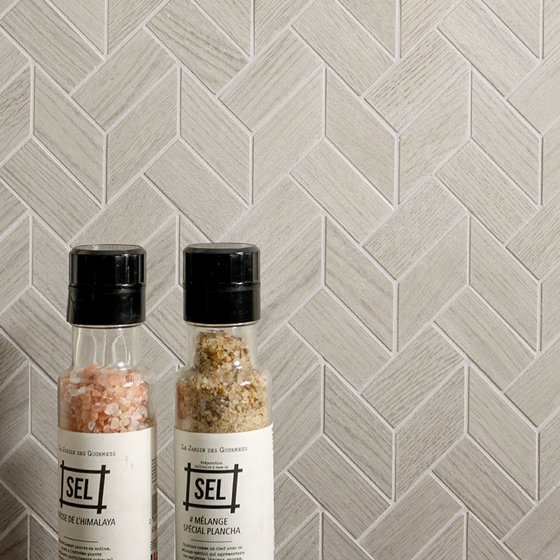 Dos botellas de especias se apoyan contra una pared de azulejos en forma de espiga de color gris claro. El texto en las botellas