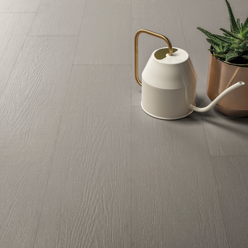Um ladrilho cinza claro com textura de madeira está no chão, acompanhado por um regador branco com alça dourada ao lado de um va