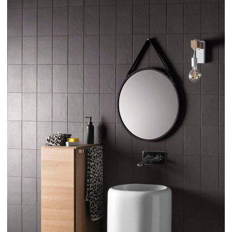 Um espelho redondo está pendurado acima de uma pia cilíndrica branca em um banheiro com paredes de azulejos escuros. Um armário 