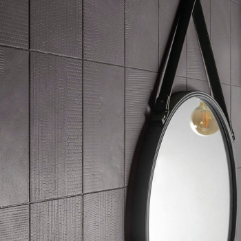 Eine strukturierte, dunkelgrau geflieste Wand spiegelt einen runden Spiegel wider, an dem ein Riemen hängt, der das warme Leucht