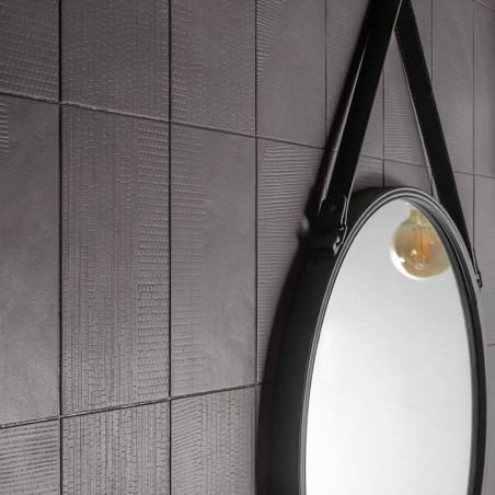 Eine strukturierte, dunkelgrau geflieste Wand spiegelt einen runden Spiegel wider, an dem ein Riemen hängt, der das warme Leucht