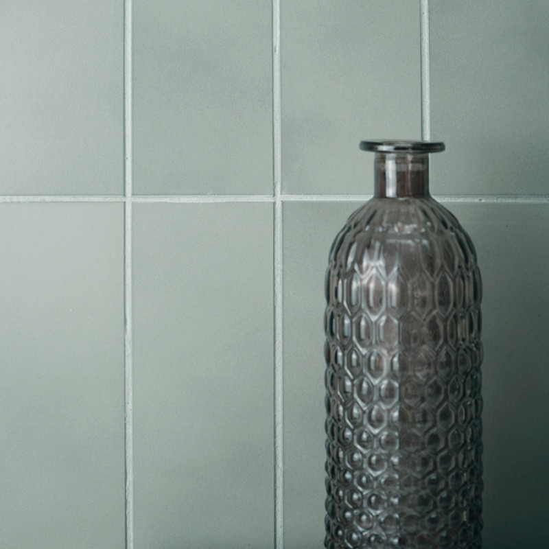 Uma garrafa de vidro alta e texturizada tem como pano de fundo azulejos quadrados cinza claro foscos, sugerindo uma estética min