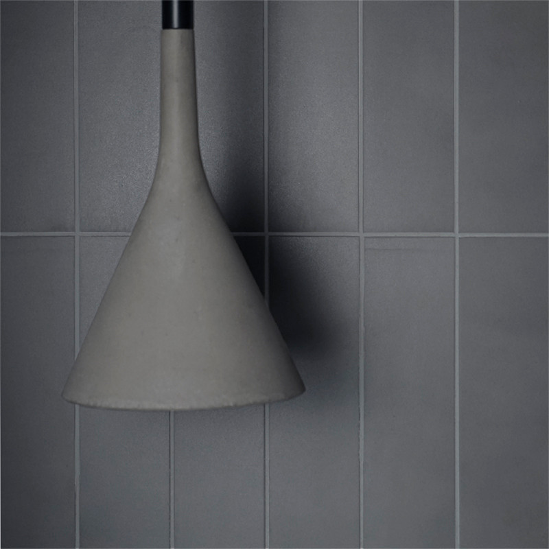 Uma luminária em forma de cone está pendurada em um fundo de azulejos cinza lisos e retangulares, criando uma estética minimalis