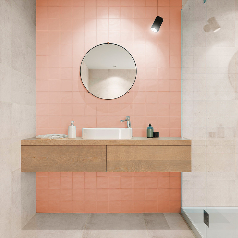 Un espejo circular cuelga sobre un tocador de madera con lavabo y artículos de tocador contra una pared de azulejos rosas en un 