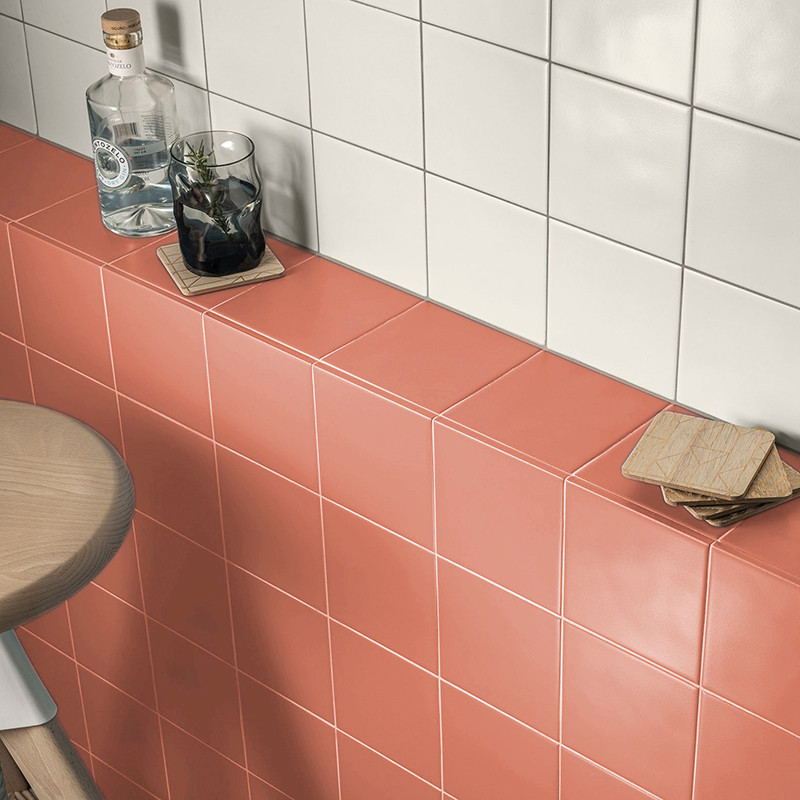 Bottles and glasses rest on coral and white tiled surfaces in a kitchen setting, with coasters nearby on a wooden stool.