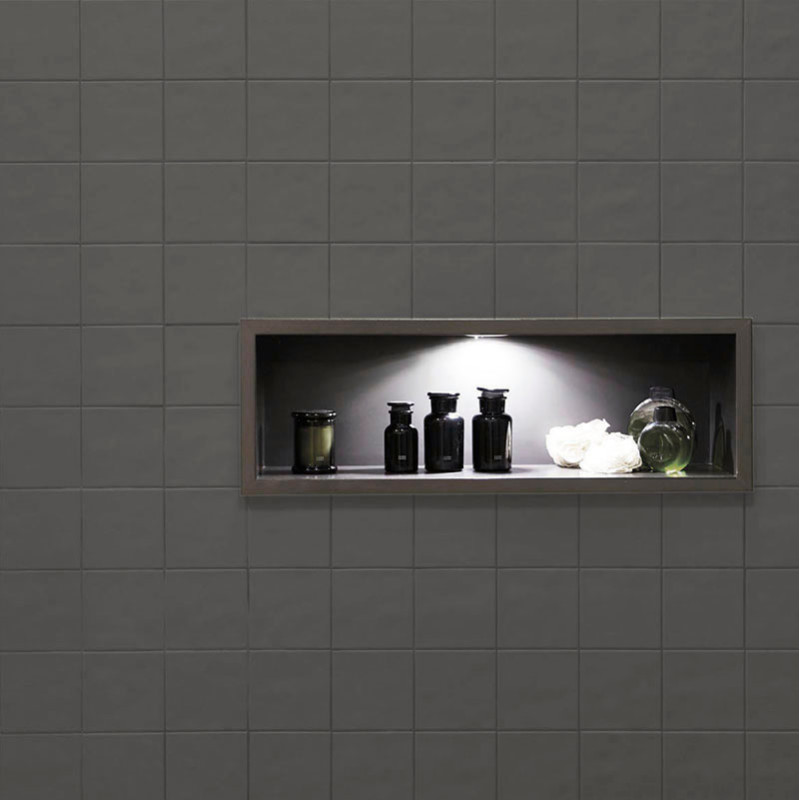Un estante empotrado con botellas negras y un jarrón verde bajo una iluminación suave se encuentra dentro de una pared de azulej