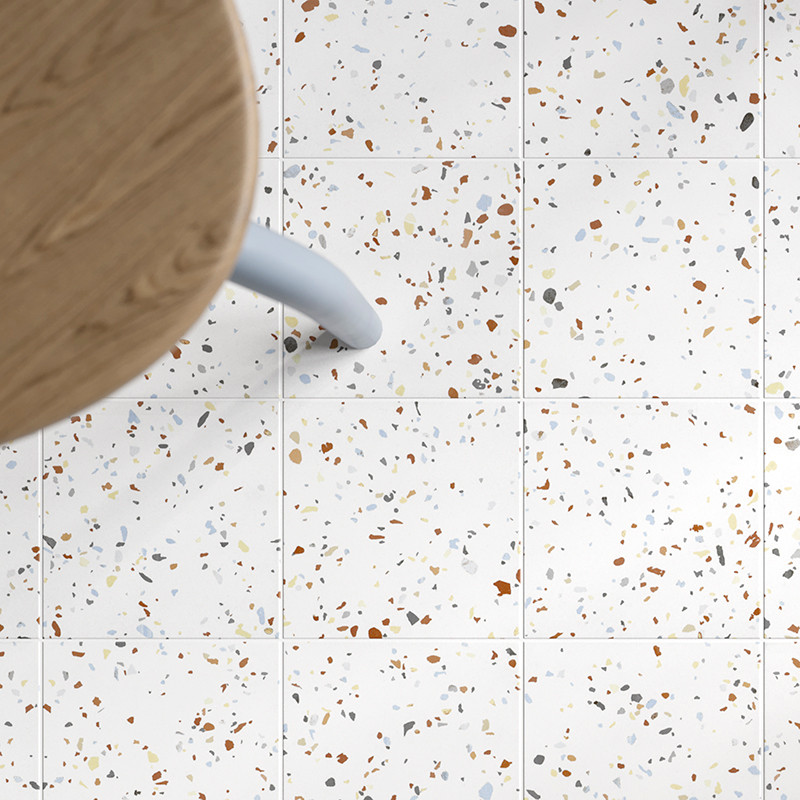 Vierkante tegels met een gespikkeld patroon van veelkleurige fragmenten liggen onder een houten tafel met een lichtblauwe poot, 