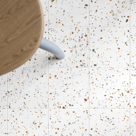 Square tiles with a speckled pattern of multicolored fragments lie beneath a wooden table with a light blue leg, creating a mode