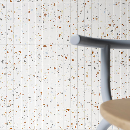 Terrazzo tiles display scattered colorful fragments on a white background. A blurred wooden seat and gray chair back are in the  Terrazzo tiles display scattered colorful fragments on a white background. A blurred wooden seat and gray chair back are in the