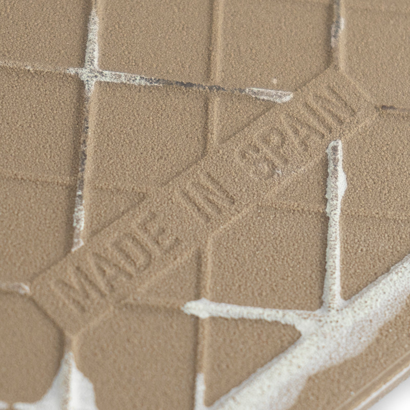 Un azulejo de cerámica marrón texturizado presenta líneas de cuadrícula en relieve y el texto `MADE IN SPAIN`, sobre un fondo li