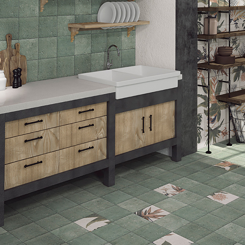 A kitchen counter features wooden drawers and a large sink, surrounded by earthy green tiles with floral patterns, in a rustic s