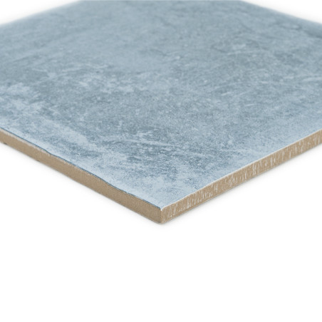 Square ceramic tile with a textured light blue surface, resting flat. The edge reveals a light brown layer.