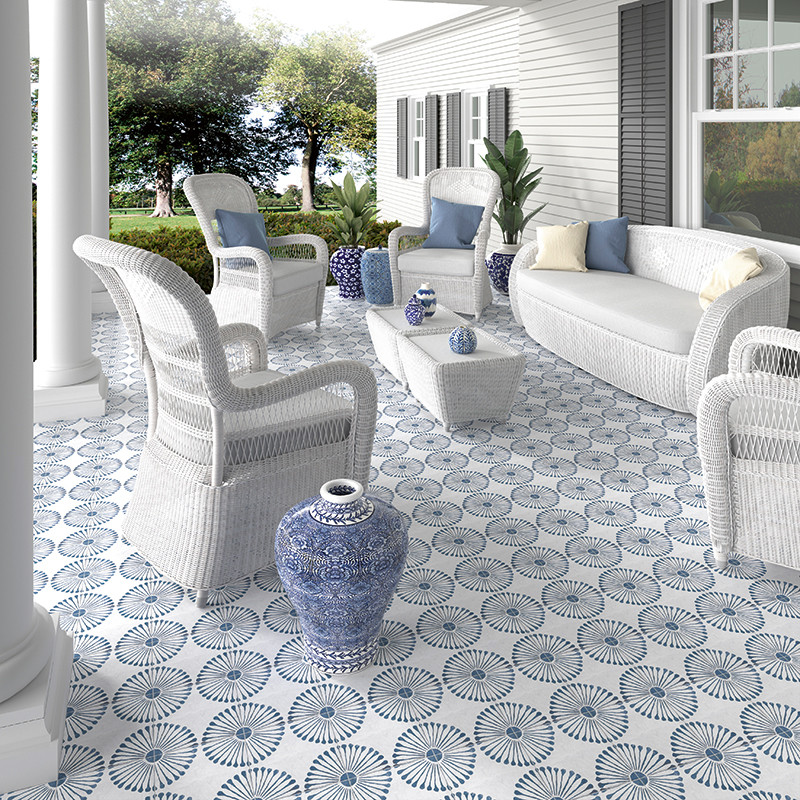 Muebles de mimbre blanco se encuentran en un patio de azulejos azules y blancos estampados. Las plantas y árboles circundantes d