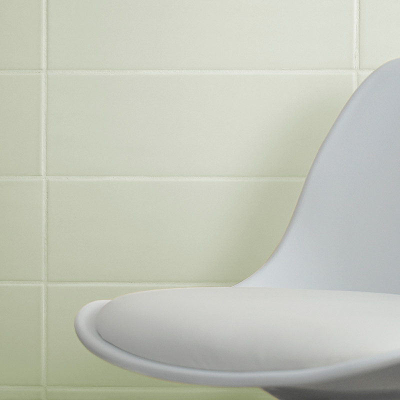 Una moderna silla blanca curvada se apoya contra una pared de azulejos lisos de color verde pálido, creando un ambiente interior