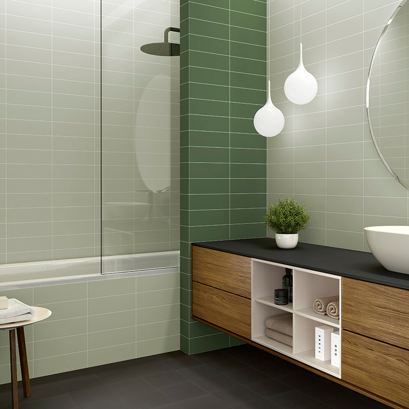Bathroom with light and dark green tiles. bathtub with glass panel. wooden vanity holding a sink, plant, and shelves beneath.