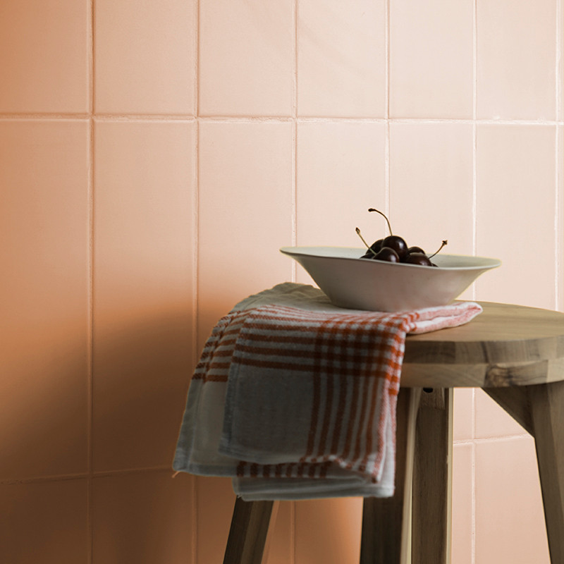 A wooden stool holds a white bowl with cherries and a red-checked cloth against a background of soft peach tiles.