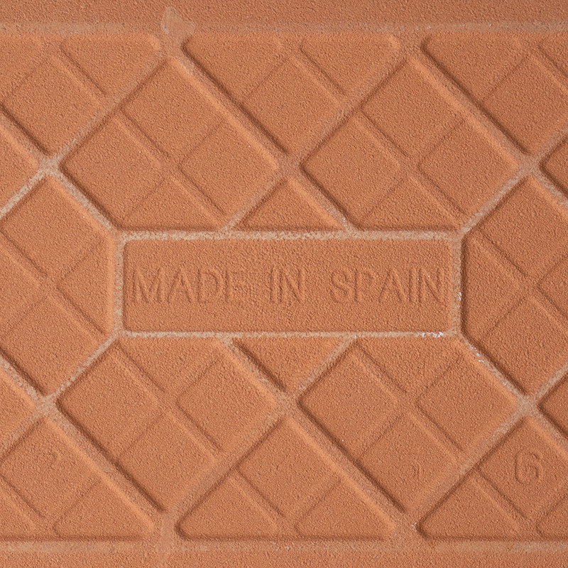 Een terracotta tegel is voorzien van de tekst `MADE IN SPAIN` in reliëf, omgeven door een geometrisch patroon van elkaar kruisen