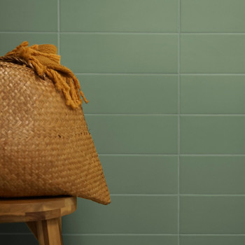 Ein geflochtener Korb mit Quasten steht auf einem Holzhocker vor einem Hintergrund aus glatten, grünen Fliesen.