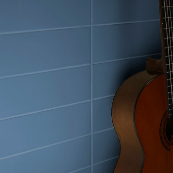 Een gitaar leunt tegen een muur die bestaat uit blauwe horizontale tegels, waarbij de snaren van de gitaar vaag zichtbaar zijn e