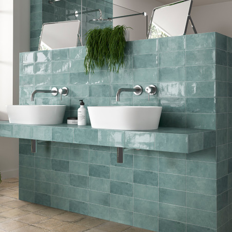 Weiße Waschbecken stehen auf einer blaugrün gefliesten Arbeitsplatte, darüber befinden sich Wasserhähne. Spiegel und eine Pflanz