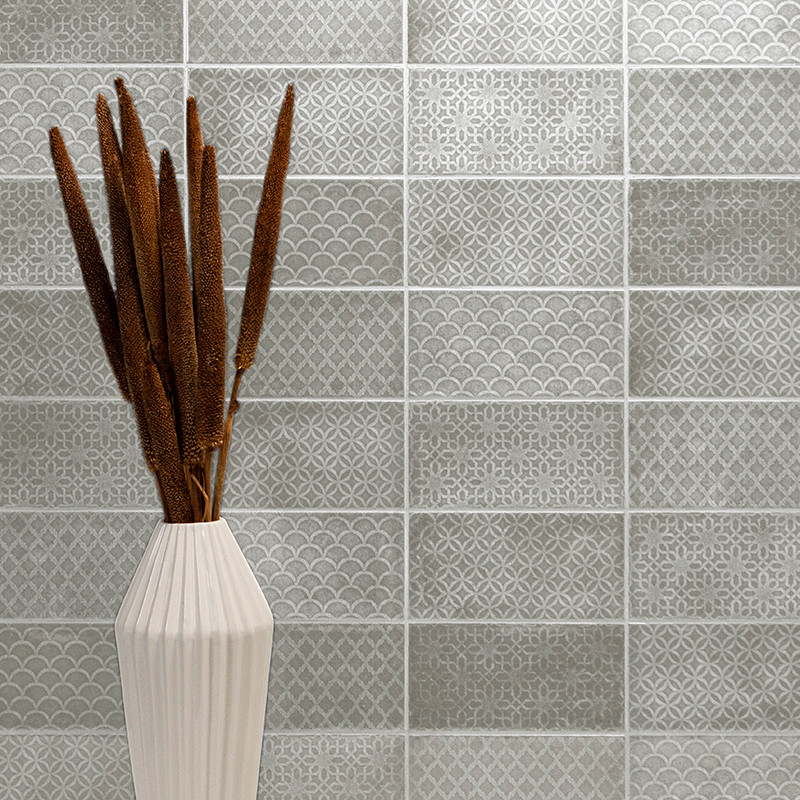 Un vase en céramique blanche contient de hautes quenouilles brunes. Derrière lui, un mur carrelé présente un motif répétitif de 