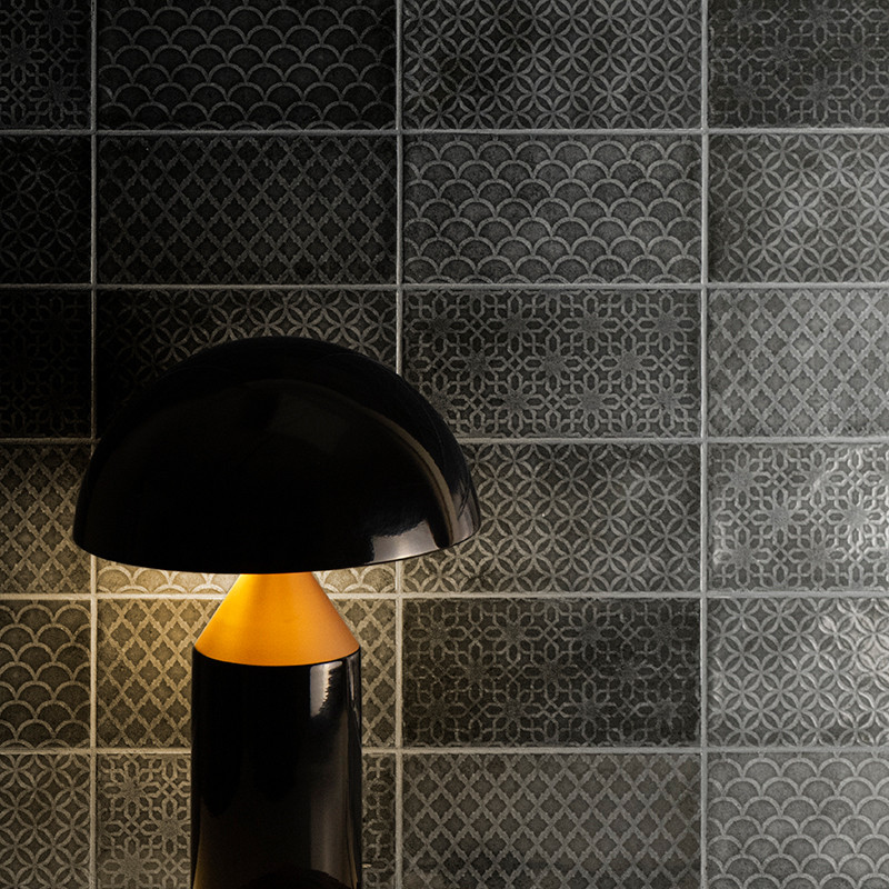 A black lamp softly illuminates intricate gray tiles with geometric and scalloped patterns in a dimly lit room.
