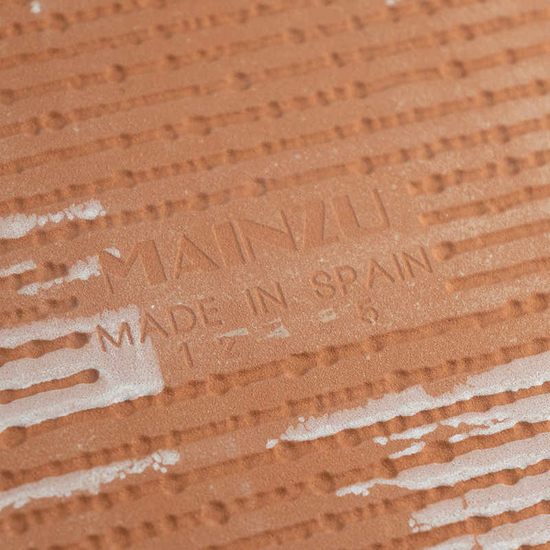 A terracotta tile, textured with raised horizontal lines and white patches, displays the text `MAINZU MADE IN SPAIN` in its cent
