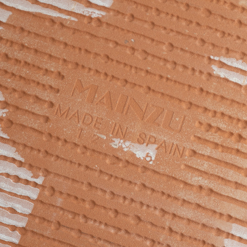 Eine orangebraune Keramikfliese, strukturiert mit linearen Mustern. Der Text „MAINZU MADE IN SPAIN“ ist in die Oberfläche eingep