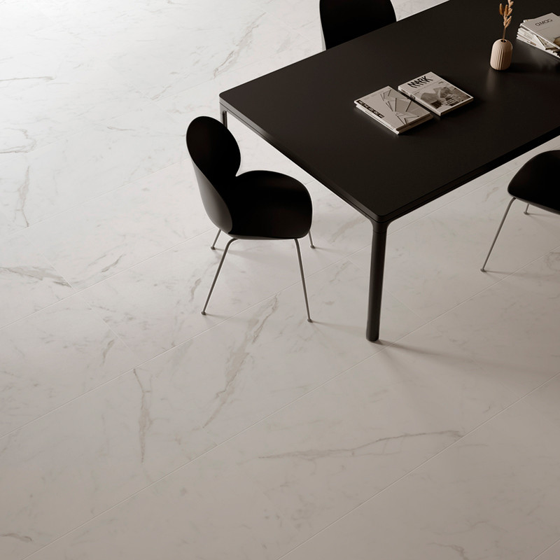 Uma elegante mesa preta com livros fica sobre um piso liso de mármore branco. Ao seu redor estão modernas cadeiras pretas com co