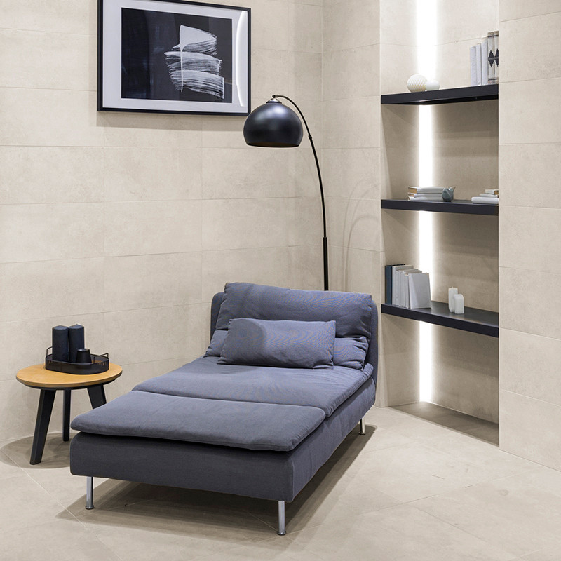 Une chaise longue grise se trouve dans une pièce minimaliste aux murs carrelés beiges. Il repose sous un lampadaire noir, à côté