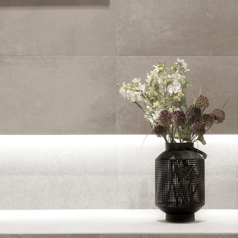 Een zwarte vaas met witte en paarse bloemen, zittend op een lichte stenen rand tegen een getextureerde beige betegelde muur.